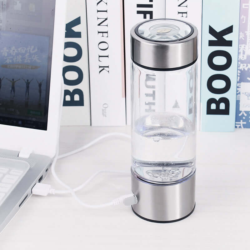 Smart Hydrogen Water Cup on desk charging via USB, standing between laptop and books, enhancing hydration with hydrogen power.