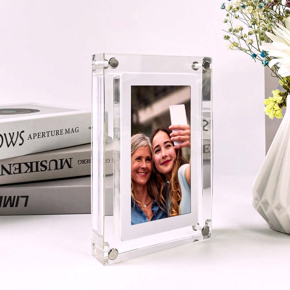 Digital picture frame acrylic video player displaying a photo of two women taking a selfie, placed on a stylish desk next to books and flowers.